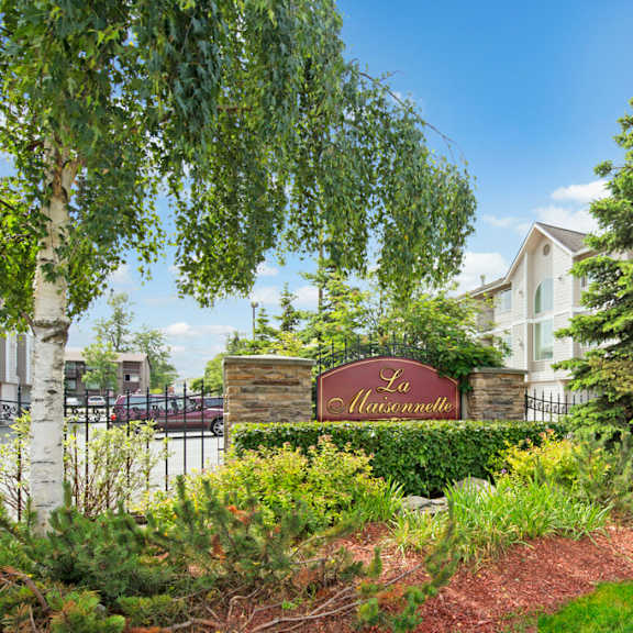lush garden with trees surrounds a sign labeled "la maisonnette." a light-colored building is in the background under a clear blue sky.