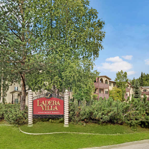 a serene apartment complex with a sign reading "ladera villa" amidst lush greenery and tall trees. the buildings are cream with arched windows. the sky is clear.
