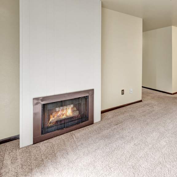 cozy living room here at ladera villas apartments with beige carpet and soft lighting. a corner fireplace adds warmth. a glimpse of the kitchen area is visible through an open doorway.