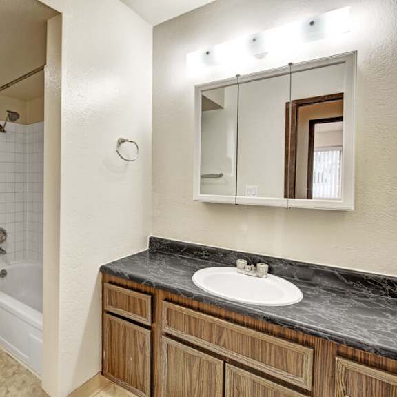 a tidy bathroom here at ladera villas apartments with a bathtub-shower combo, white tiles, and a white toilet. a wooden vanity with a marble countertop sits beneath a lit mirror.