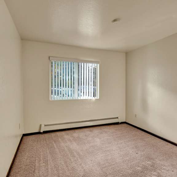 bedroom here at ladera villas apartments with beige carpet and cream walls, lit by sunlight through vertical blinds on a window. the tone is warm and inviting.