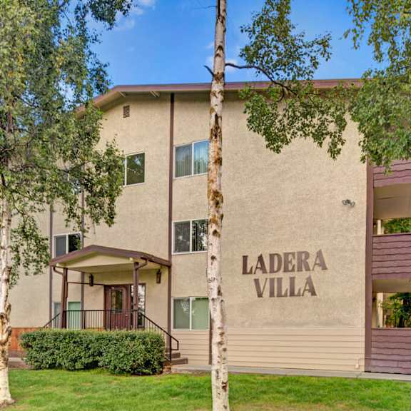 three-story beige ladera villa apartment building labeled "ladera villa" with brown accents. surrounded by lush green trees and grass under a clear blue sky.