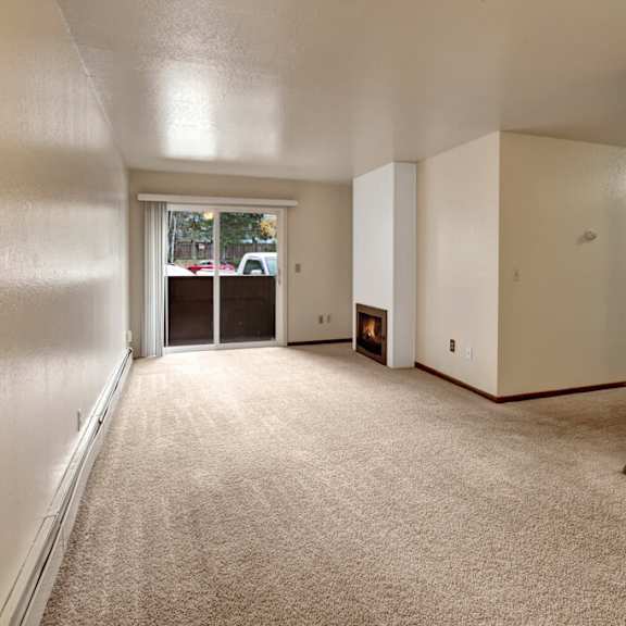 a spacious, empty living room here at ladera villas apartments with beige carpet and white walls. a lit fireplace adds warmth. a sliding glass door opens to a patio, revealing parked cars outside.