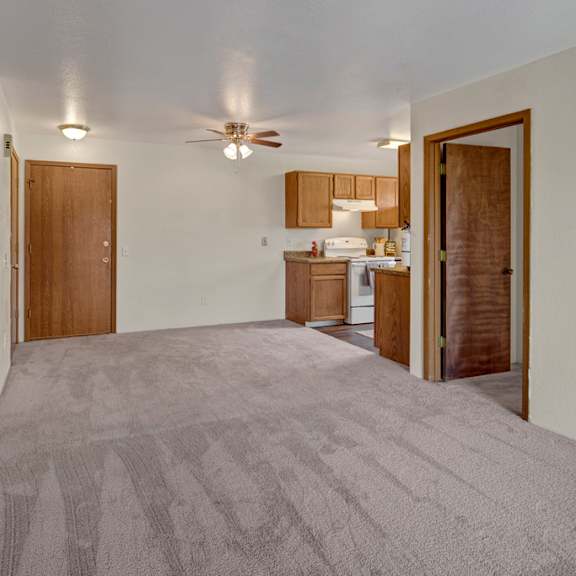 spacious living area here at legacy west apartments with beige carpet and neutral walls, connected to a kitchen with wooden cabinets and white appliances. ceiling fan above adds warmth.