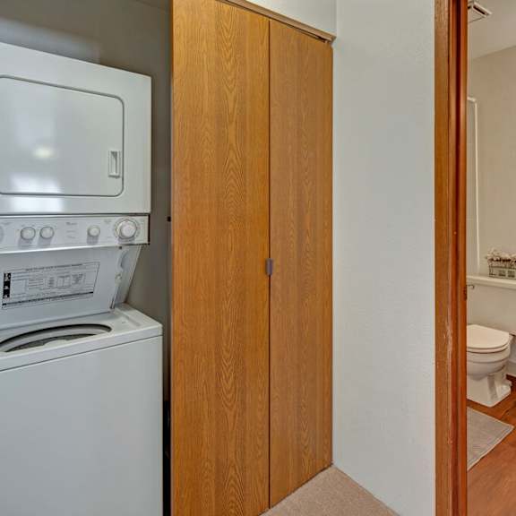 laundry area here at legacy west apartments with a stacked washer and dryer next to wooden cabinets. adjacent bathroom with sink, mirror, toilet, and warm lighting; cozy atmosphere.
