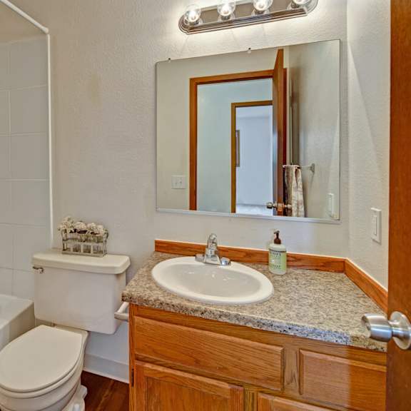 small bathroom here at legacy west apartments with white tiled shower-tub combo, beige curtain, wooden vanity, granite counter, oval sink, mirror, and chrome light fixture.