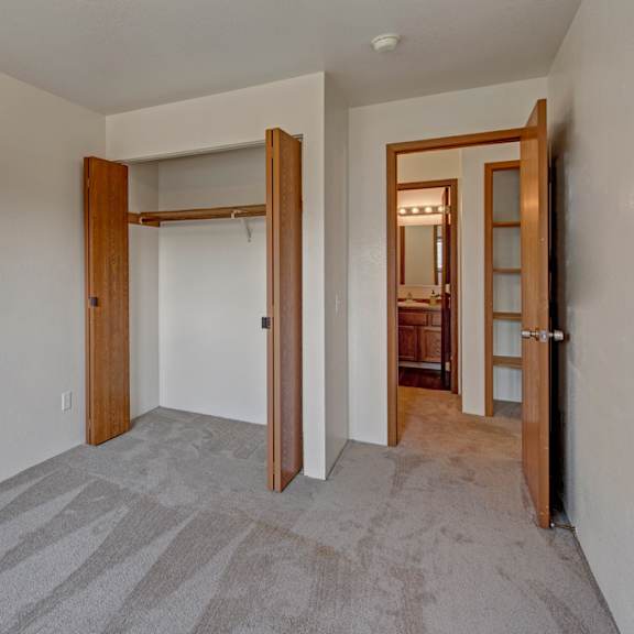 empty bedroom here at legacy west apartments with light gray carpet, open wooden closet doors, and an adjoining bathroom door. neutral walls and a tidy ambiance.