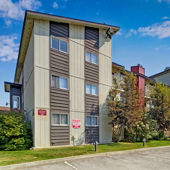 three-story legacy west apartment building with tan and brown siding, surrounded by trees and grass. clear blue sky, empty parking lot in the foreground.