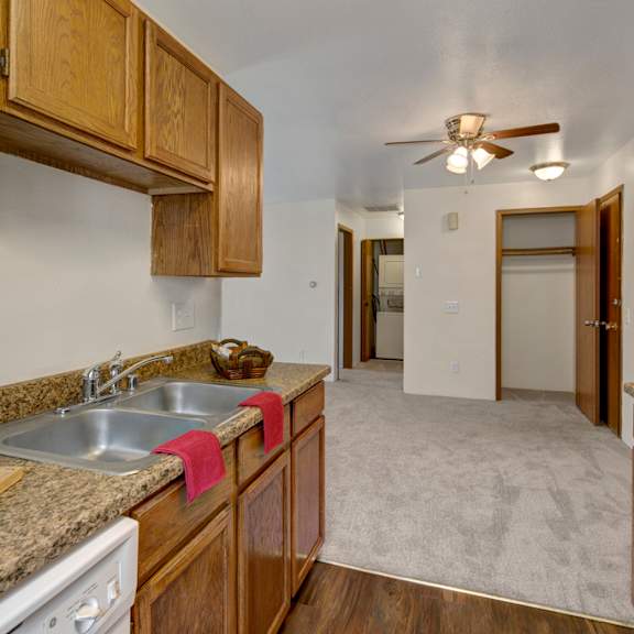kitchen here at legacy west apartments with wood cabinets and granite countertops, double sink with red towel, leading to carpeted hall. ceiling fan and open closet visible. cozy ambiance.