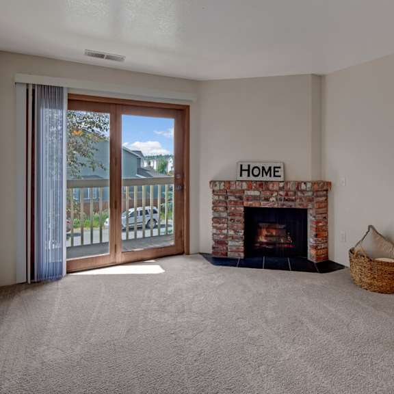 empty living room here at legacy west apartments with a cozy brick fireplace, sign reading "home," and a wicker basket on plush carpet. sunlit balcony doors reveal trees outside.