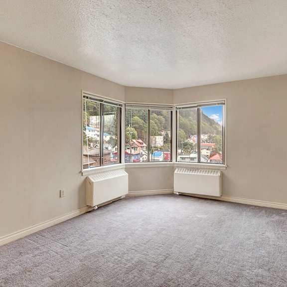 living room here at mendenhall tower apartments with beige walls and gray carpet, featuring a bay window. the window offers a view of colorful houses and lush green hills under a blue sky.