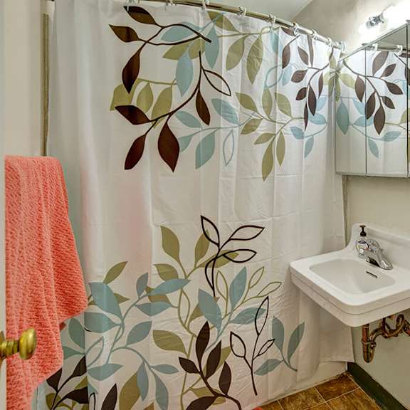 bathroom here at mendenhall tower apartements with a leafy pattern shower curtain, white sink, and mirror. a coral towel hangs nearby, creating a calm, fresh atmosphere.