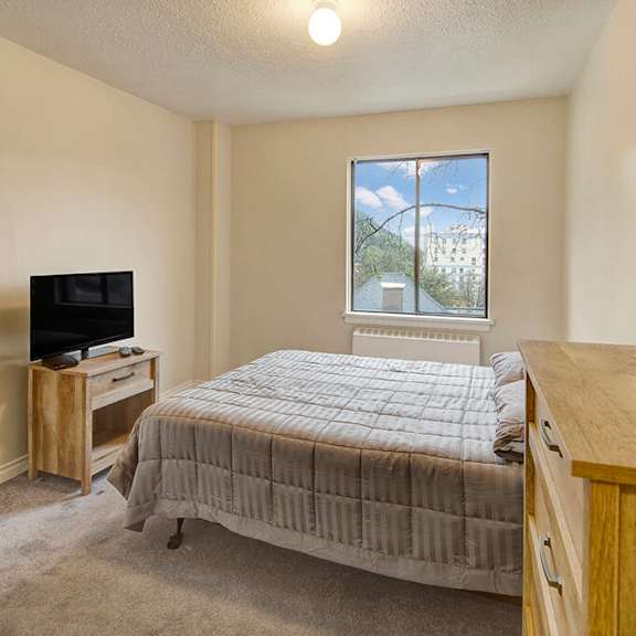 a cozy bedroom here at mendenhall tower apartment featuring a wooden bed with a gray quilt, a small tv on a stand, a dresser with a teal lamp, and a window with a view of trees.