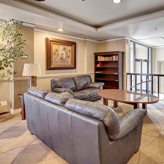 cozy lounge area here at mendenhall tower apartments with two gray leather sofas facing a round, wooden coffee table. a bookshelf, framed artwork, and large potted plants enhance the warm atmosphere.