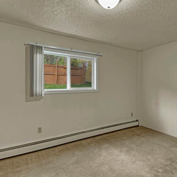 empty bedroom here at montclair apartments with beige carpet and off-white walls, featuring a single window with vertical blinds and a view of a wooden fence and greenery outside. neutral and calm atmosphere.