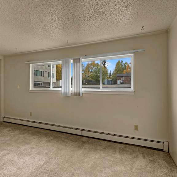 empty bedroom here at montclair apartments with light beige walls and carpet, large window with vertical blinds showing trees and a building outside. calm, neutral atmosphere.