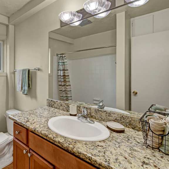 a modern bathroom here at montclair apartments with a granite countertop and sink, adjacent to a toilet. towels hang by a frosted window, and soft, warm lighting creates a cozy ambiance.