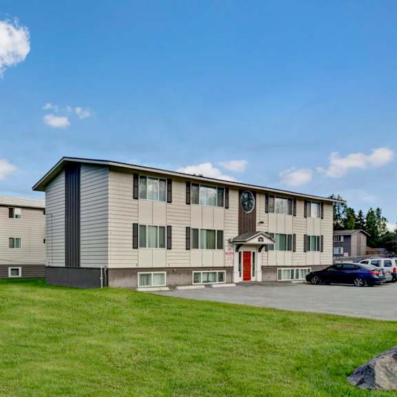 two-story montclair apartment building with white siding and red door set against a blue sky with fluffy clouds. surrounding green lawn adds a serene touch.