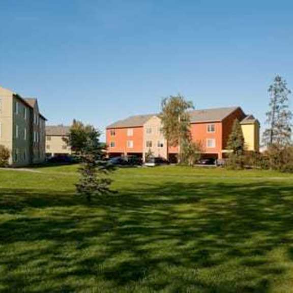 sunny park scene here at brighton park apartments with green lawn in foreground. two residential buildings, one yellow and one red, are surrounded by trees under a clear blue sky.