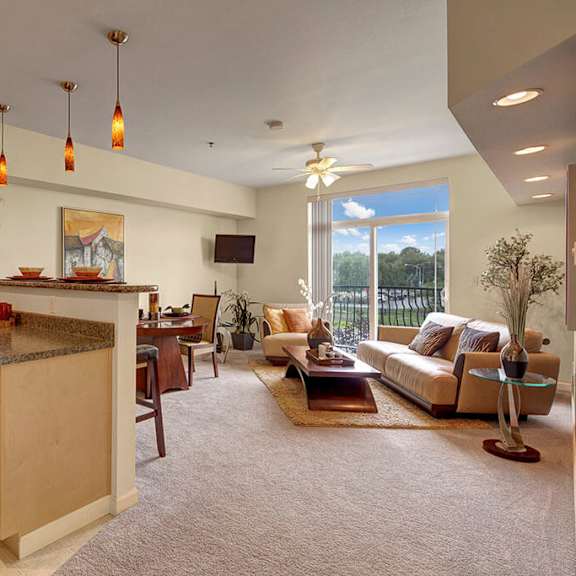 spacious open-plan living room and kitchen here at park plaza 2 apartments. light wood cabinets, pendant lights, cozy seating, large window with greenery view, warm, inviting atmosphere.