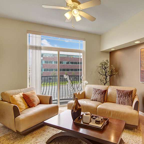 bright modern living room here at park plaza 2 apartments with beige sofas, abstract painting, ceiling fan, and large window showing a view of an office building. cozy and inviting.