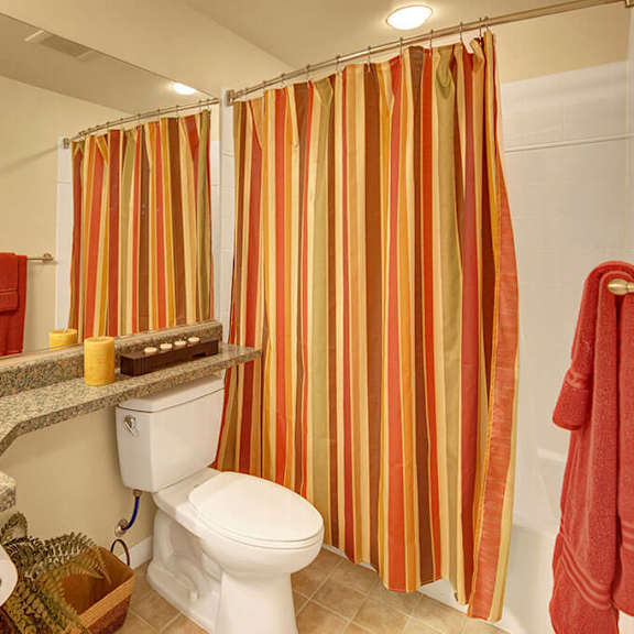 bathroom here at park plaza 2 apartments with red and yellow striped shower curtain, granite countertop, white toilet, and a mirror. red towels and candles add warmth and coziness.