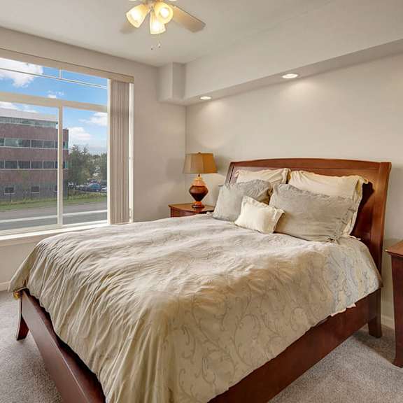 modern bedroom here at park plaza 2 apartments with a large window view, featuring a wooden bed with beige and gray bedding, side tables, a tall vase, and a warm, inviting ambiance.