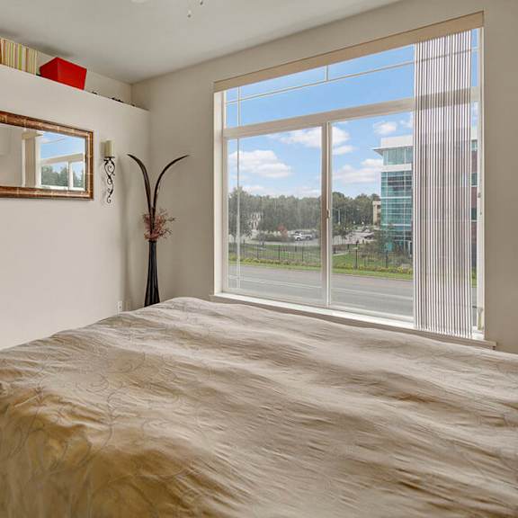 spacious bedroom here at park plaza 2 apartments with large window offering a view of trees and buildings. elegant wall mirror and vase add sophistication. calm and cozy feel.