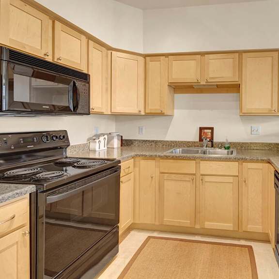 a cozy kitchen here at park plaza 2 apartments features light wood cabinets, granite countertops, a black stove, and a microwave. red mugs and a vase add warm touches to the space.