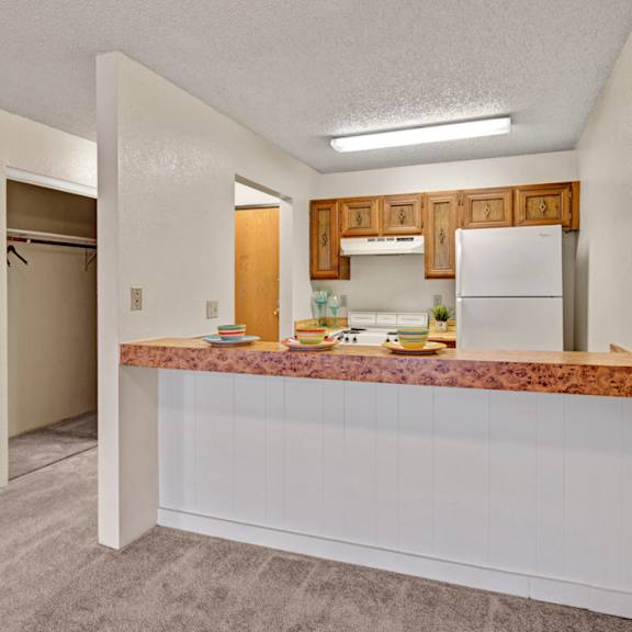 compact kitchen here at park plaza apartments with a wood-accented breakfast bar, set with colorful plates. white appliances, wood cabinets, and an open closet create a cozy, inviting feel.