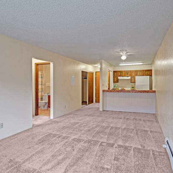 spacious empty living room here at park plaza apartments with beige carpet and cream walls, leading to a kitchen with brown cabinets and a bathroom visible on the left. cozy and inviting.