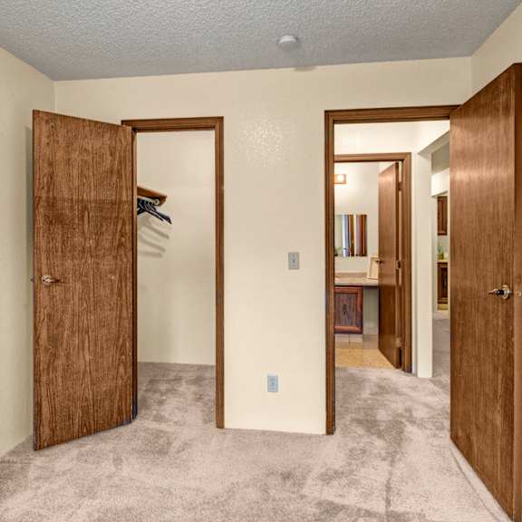 a small bedroom here at park plaza apartments with beige walls and carpet. two open wooden doors show a closet with hangers on the left and a bathroom with a mirror on the right.