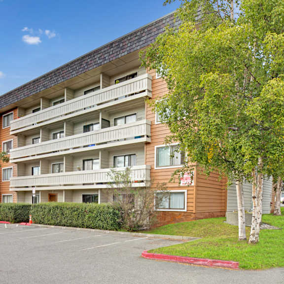 four-story park plaza apartment building with white balconies, surrounded by green trees and a parking lot with several cars, under a clear blue sky.