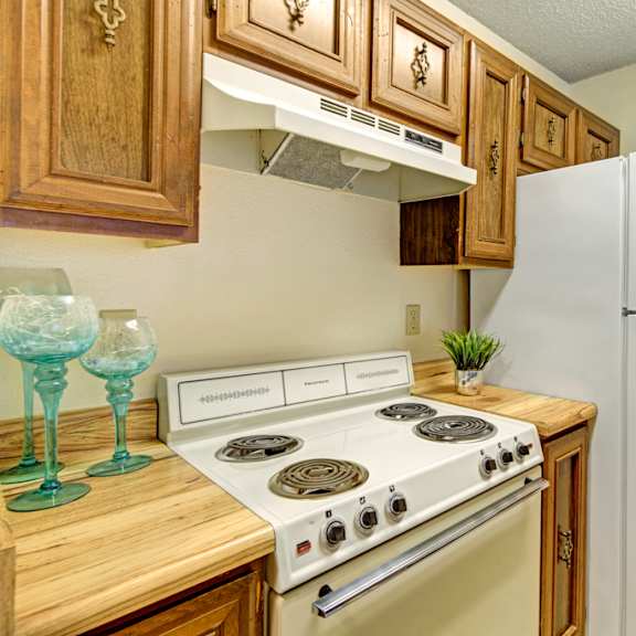 compact kitchen here at park plaza apartments with white appliances, including a stove and refrigerator. wooden cabinets above, light countertop with teal glasses and a potted plant. cozy vibe.