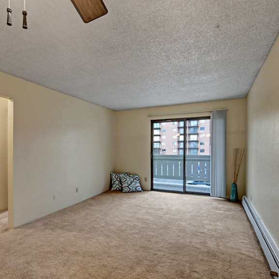 spacious, empty living room here at park plaza apartments with beige walls and carpet, ceiling fan, and sliding glass door opening to a balcony. minimalist decor includes a cushion and a vase.