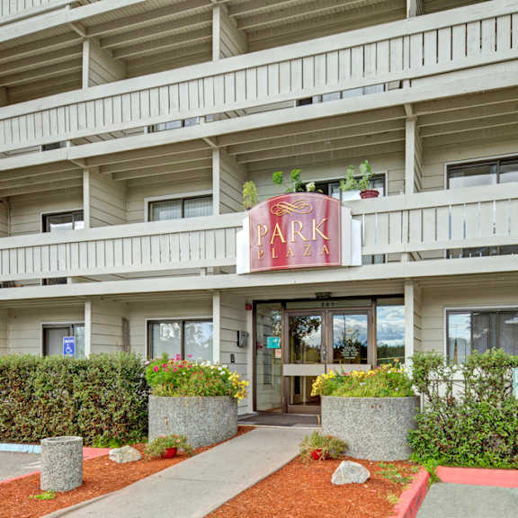 facade of a beige building with a "park plaza" sign, featuring balconies and entrance surrounded by greenery and colorful flower planters.