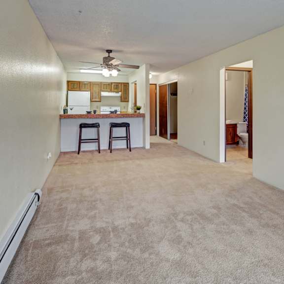 long carpeted living room here at park plaza apartments leading to an open kitchen with wooden cabinets and a ceiling fan. two bar stools at a counter. neutral tones, calming ambiance.