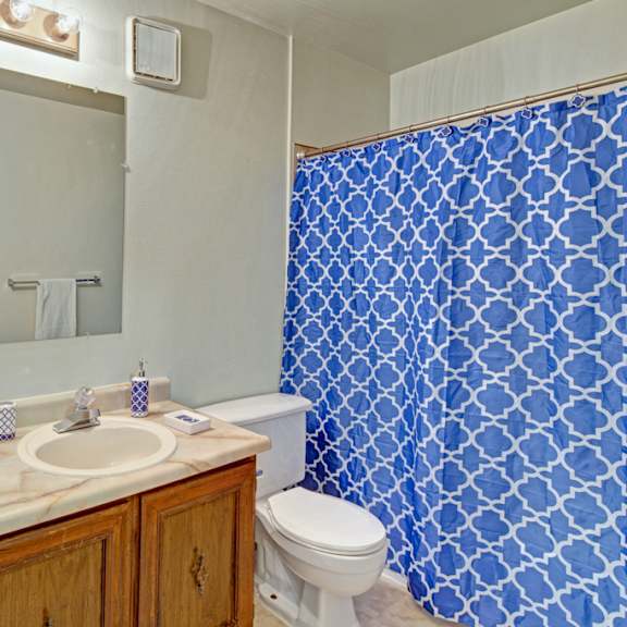 bathroom here at park plaza apartments with marble countertop and wood cabinets, blue geometric shower curtain, large mirror, framed beach photo, and a clean, bright atmosphere.