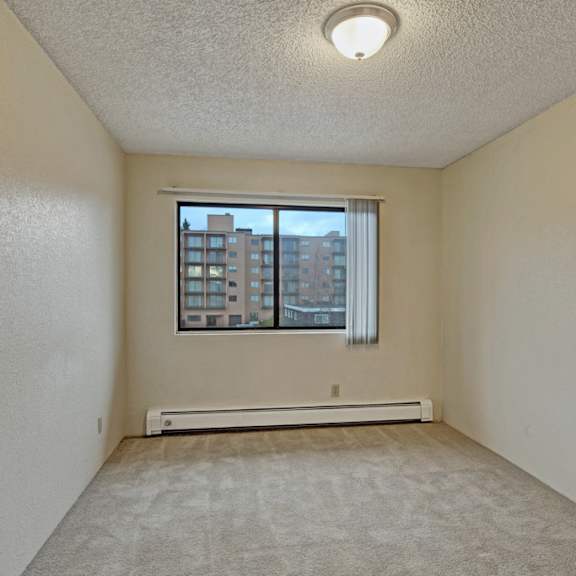 bedroom here at park plaza apartments with beige walls and carpet is lit by a ceiling fixture. a window with vertical blinds reveals a view of nearby buildings, creating a calm atmosphere.