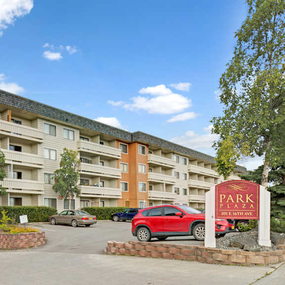four-story apartment building with beige and orange siding under a bright blue sky. cars are parked in front with a "park plaza" sign visible.