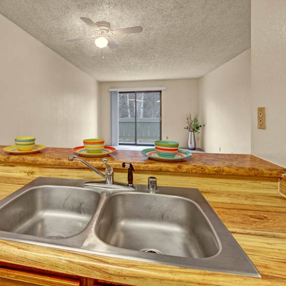 cozy kitchen here at park plaza apartments with a wooden countertop and double sink, set with colorful plates and bowls. a ceiling fan and distant sliding doors enhance the room's brightness.