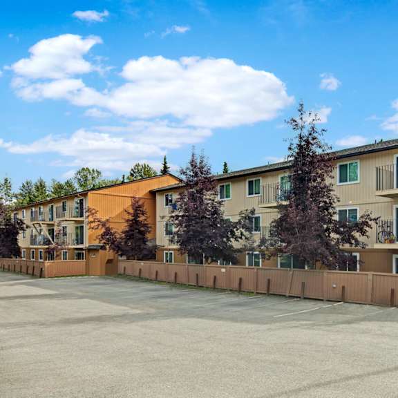 three-story beige quiet creek apartment building with brown trim, surrounded by trees and enclosed by a wooden fence, under a bright blue sky with scattered clouds.