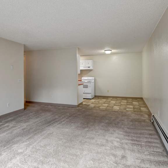 empty apartment here at quiet creek apartments with beige carpeted living area, leading to a small kitchen with white appliances and linoleum floor. bright, clean, and minimalist.