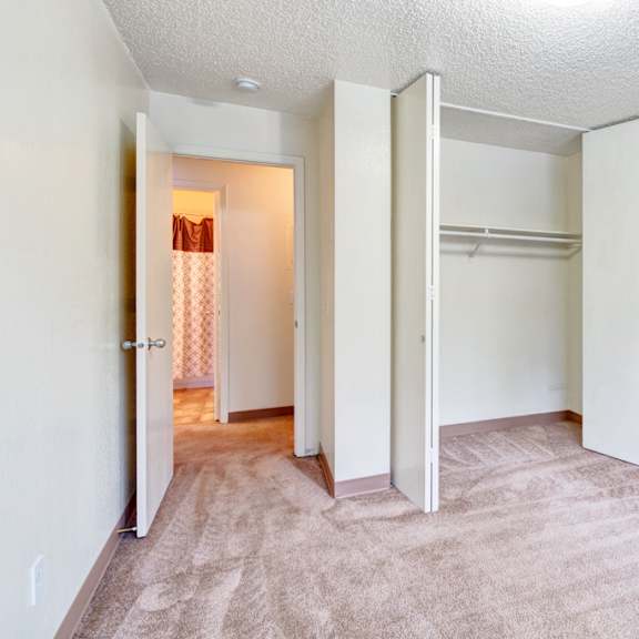 empty bedroom here at quiet creek apartments with beige carpet and white walls, featuring an open closet with shelves. a door leads to a lit hallway with a patterned shower curtain.