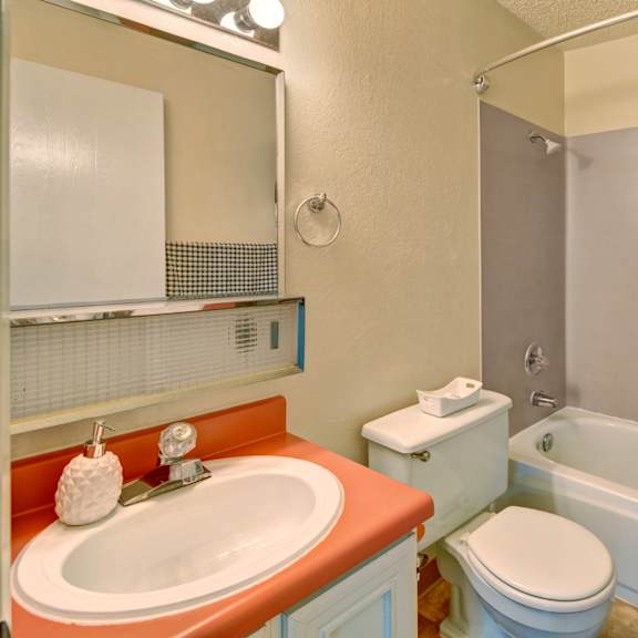 compact bathroom here at quiet creek apartments with red countertop, white sink, and decorative soap dispenser. white toilet next to a tub with a patterned shower curtain. bright lighting conveys a clean and cozy atmosphere.