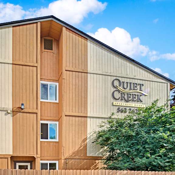 apartment building with wood paneling in beige and tan. "quiet creek apartment homes" sign on the right. trees and a wooden fence in the foreground. blue sky above.