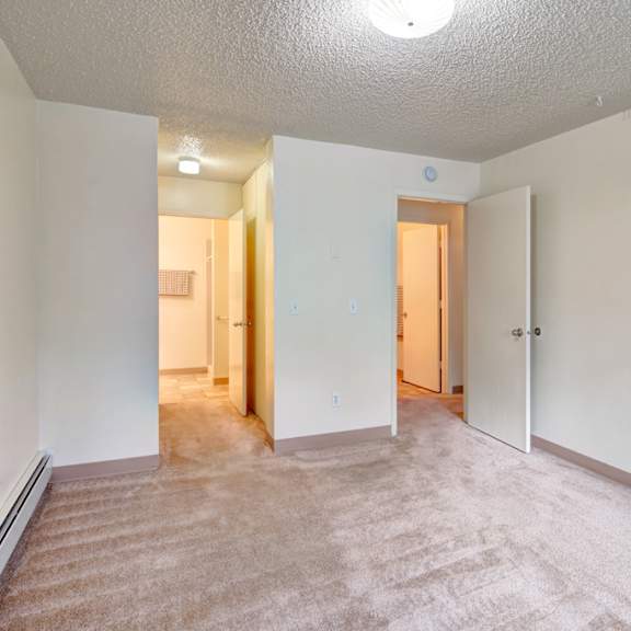 empty bedroom here at quiet creek apartments with beige carpet and off-white walls. an open door reveals a lit hallway. the room is minimalistic and calm, evoking a neutral tone.