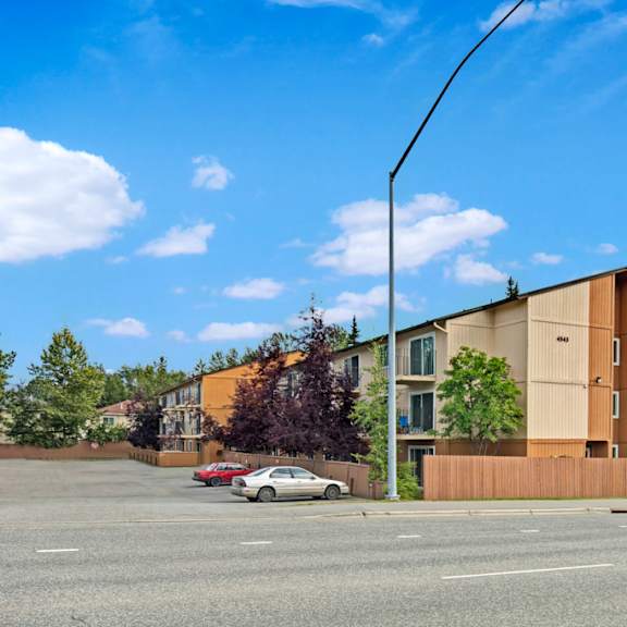 three-story apartment building with "quiet creek" sign, surrounded by trees, under a blue sky with clouds. two cars parked in front on a paved lot.