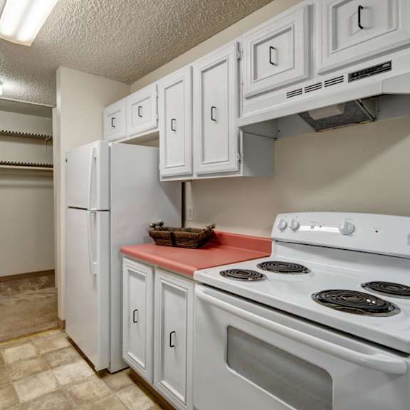 narrow kitchen here at quiet creek apartments with white cabinets, red countertops, and a white stove. a white refrigerator stands beside an open pantry with empty shelves. bright lighting.