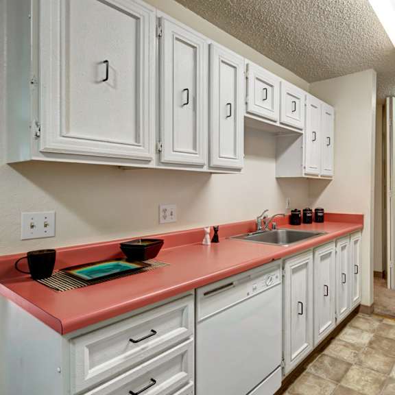 narrow kitchen here at quiet creek apartments with coral countertops, white cabinets, stove, and fridge. bright lighting, clean, organized, with a modern yet cozy feel.